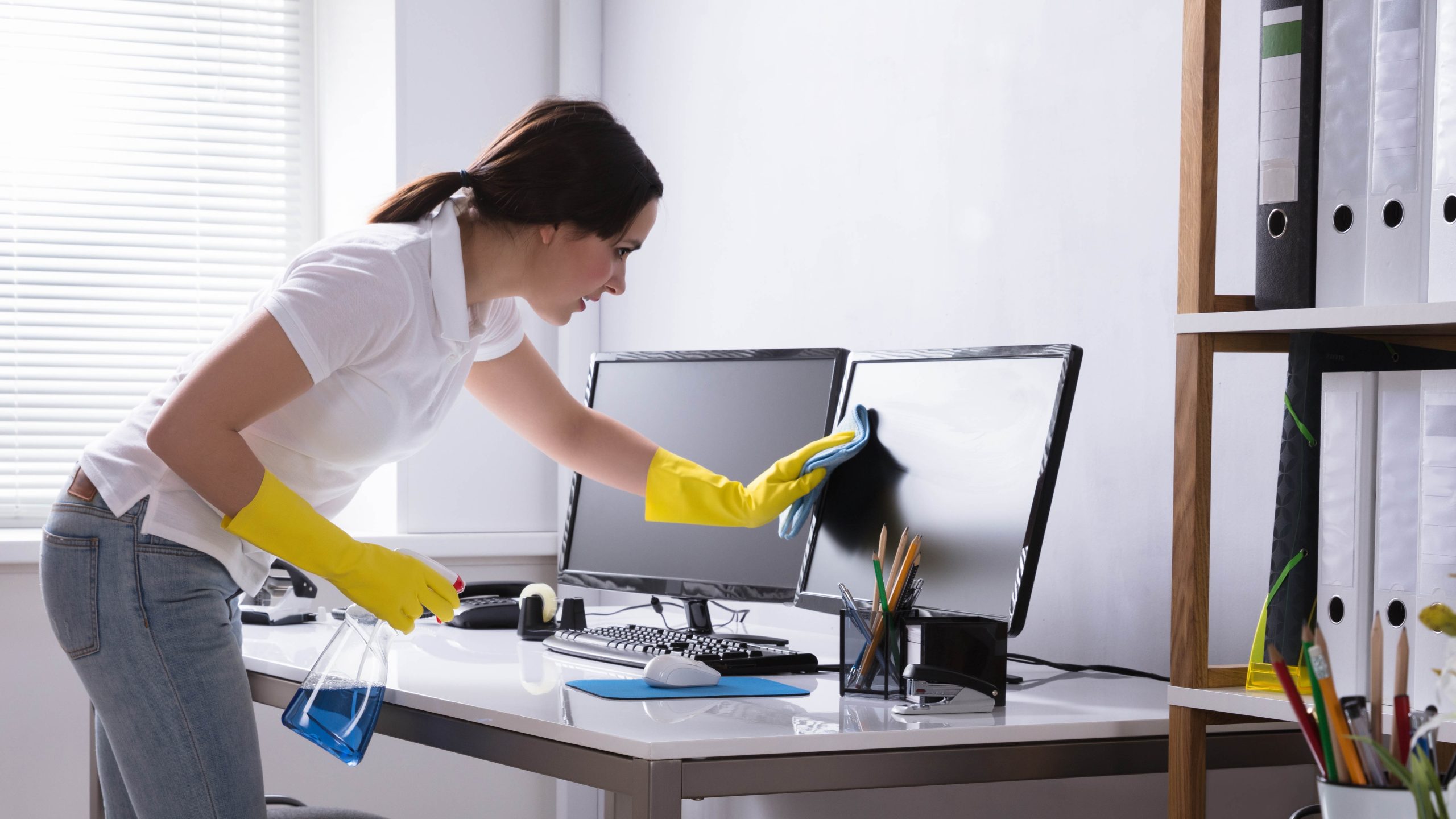 Young Woman Cleaning Computer Monitor With Rag In Office